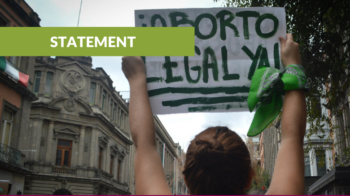 Statement. A women wearing a green bandana and holding up a sign that reads 'legal abortion now' in Spanish.