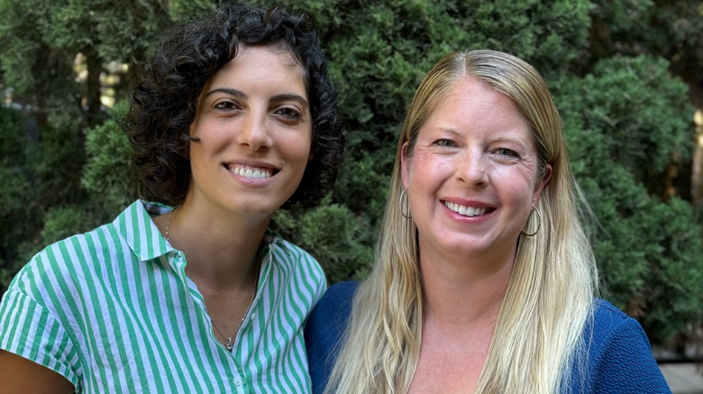 Rouba and Lisa smiling at the camera. Rouba has brown curly hair and a striped blouse. Lisa has long blonde hair and a blue dress.